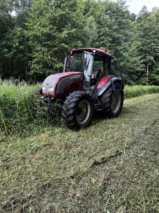 Czerwony traktor Valtra z otwartymi drzwiami na skraju lasu, świeżo skoszona trawa widoczna na ziemi.