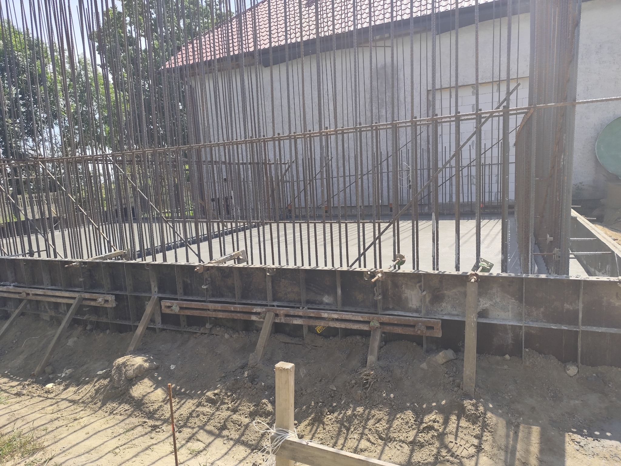 Zbrojenie fundamentów domu jednorodzinnego z widocznymi prętami stalowymi i szalunkiem, przygotowane do zalania betonem, w tle budynek z czerwonym dachem.