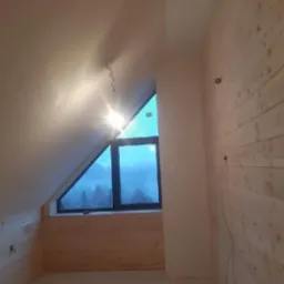 Poddasze z drewnianą boazerią i oknem w kształcie trójkąta, widok na zalesione wzgórza w oddali. Surowe wykończenie, widoczne kable. Oświetlenie górne.