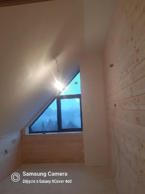 Poddasze z drewnianą boazerią i oknem w kształcie trójkąta, widok na zalesione wzgórza w oddali. Surowe wykończenie, widoczne kable. Oświetlenie górne.