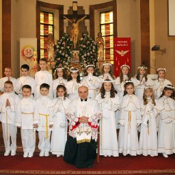 STUDIO ART.FRANKO SPÓŁKA Z OGRANICZONĄ ODPOWIEDZIALNOŚCIĄ - Grupa dzieci w białych strojach komunijnych wraz z księdzem na tle ołtarza w kościele podczas uroczystości Pierwszej Komunii Świętej.