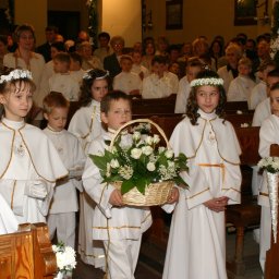 STUDIO ART.FRANKO SPÓŁKA Z OGRANICZONĄ ODPOWIEDZIALNOŚCIĄ - Grupa dzieci w białych strojach komunijnych, dziewczynki w wiankach, chłopiec niesie koszyk z białymi różami, w tle zgromadzeni w kościele.