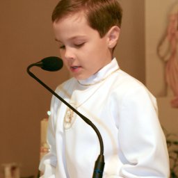 STUDIO ART.FRANKO SPÓŁKA Z OGRANICZONĄ ODPOWIEDZIALNOŚCIĄ - Chłopiec w białej albie z haftowanym symbolem IHS, czytający tekst przy mikrofonie podczas uroczystości religijnej.