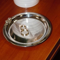 STUDIO ART.FRANKO SPÓŁKA Z OGRANICZONĄ ODPOWIEDZIALNOŚCIĄ - Dwa złote pierścionki ślubne leżące na srebrnej tacy z dekoracyjnym ornamentem, obok biały wazon z materiałową ozdobą na drewnianym stole.