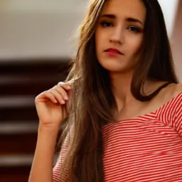 Portret młodej kobiety w bluzce w czerwono-białe paski, z ręką uniesioną do twarzy, w naturalnym oświetleniu.