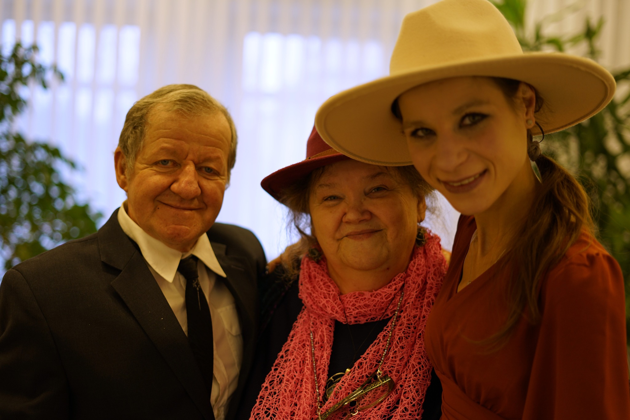 Portret trzech osób: eleganckiego mężczyzny w garniturze, kobiety w kapeluszu i różowym szalu oraz młodej kobiety w sukience i kapeluszu, uśmiechniętych i pozujących do zdjęcia.