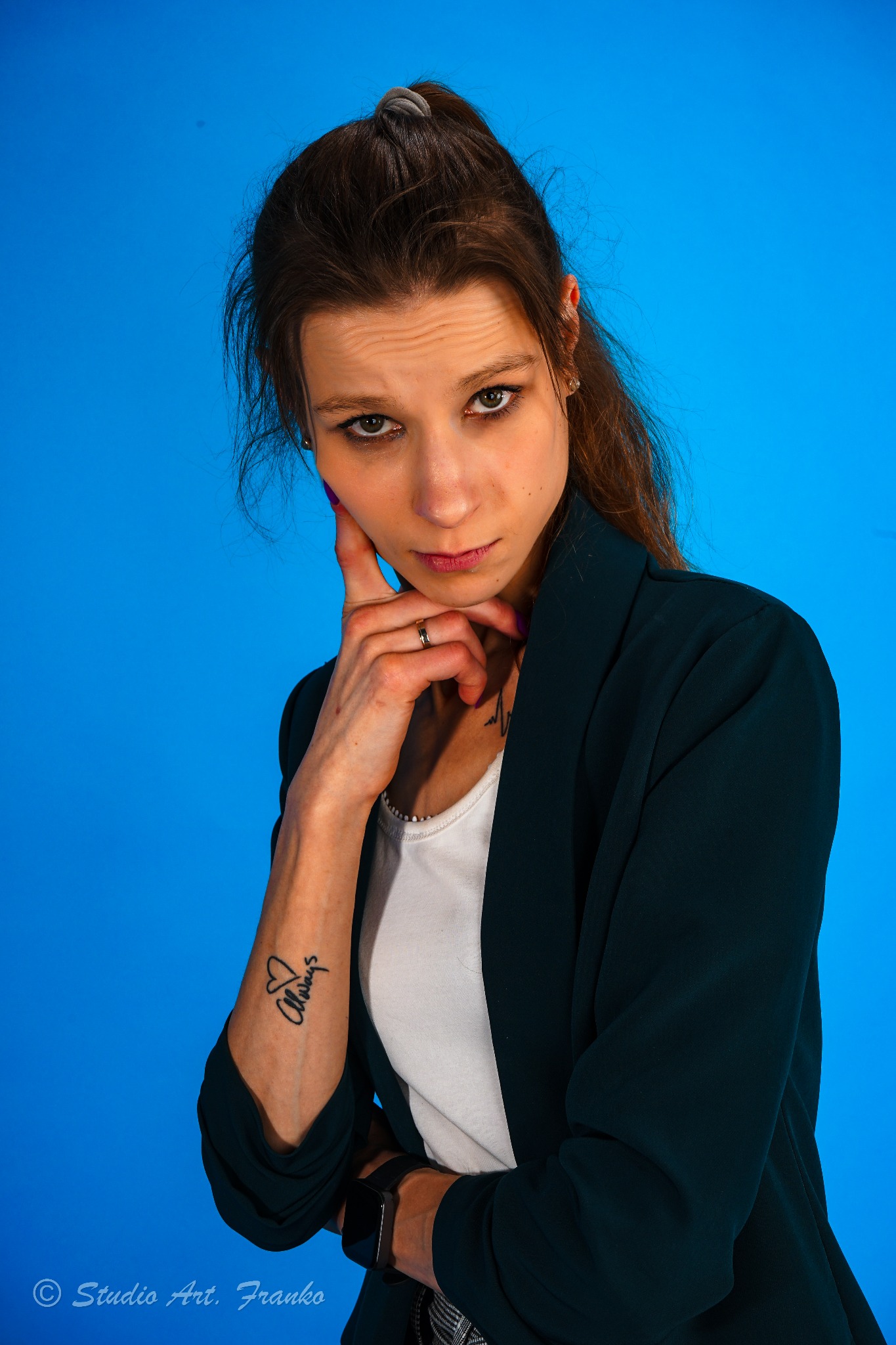 Portret kobiety w ciemnozielonej marynarce na jednolitym niebieskim tle, z ręką podpartą pod brodą, widoczny tatuaż na przedramieniu i pierścionek na palcu.