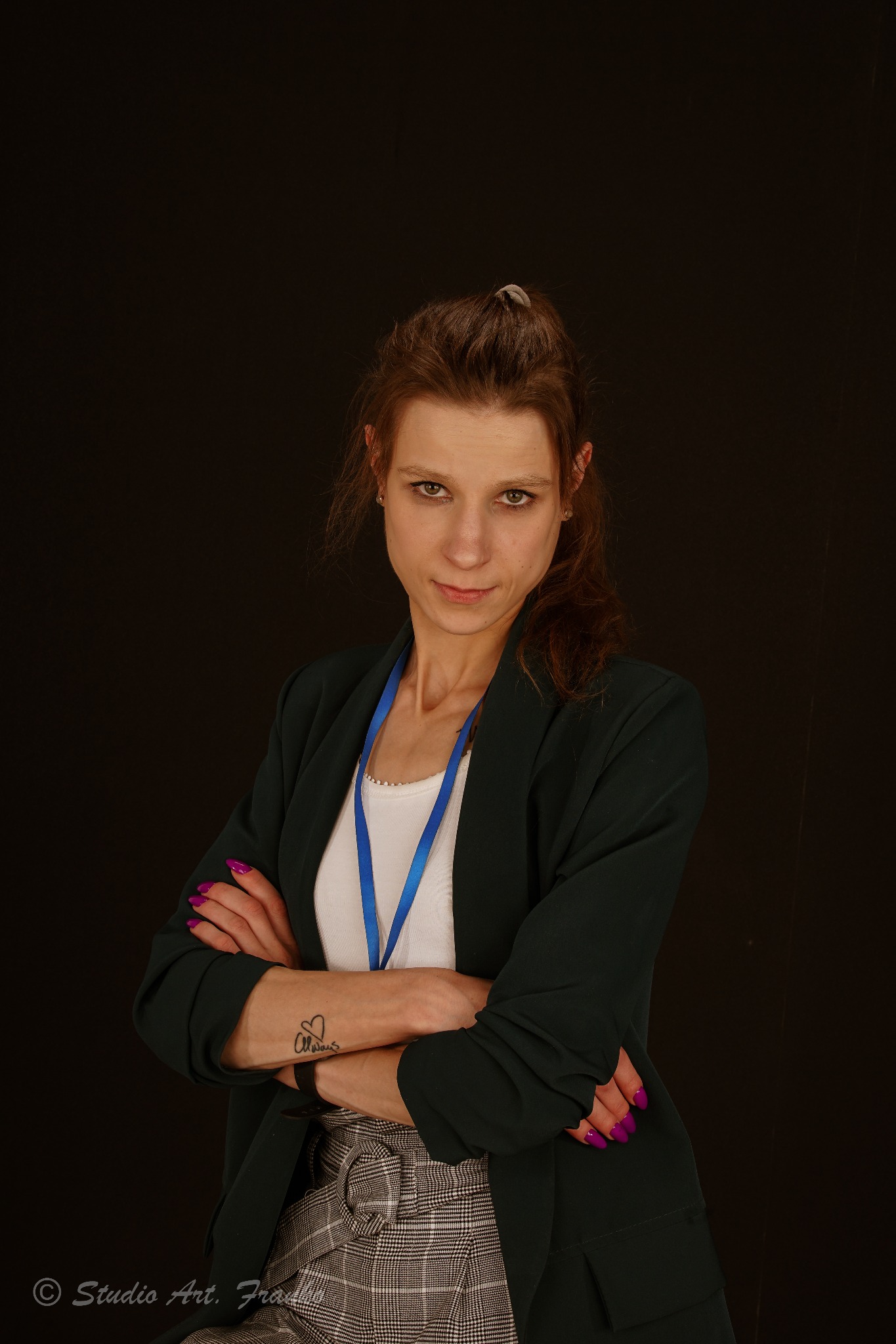 Portret kobiety z brązowymi włosami spiętymi gumką, ubrana w zieloną marynarkę i spódnicę w kratę, z identyfikatorem na szyi i tatuażem na przedramieniu, pozująca na ciemnym tle.