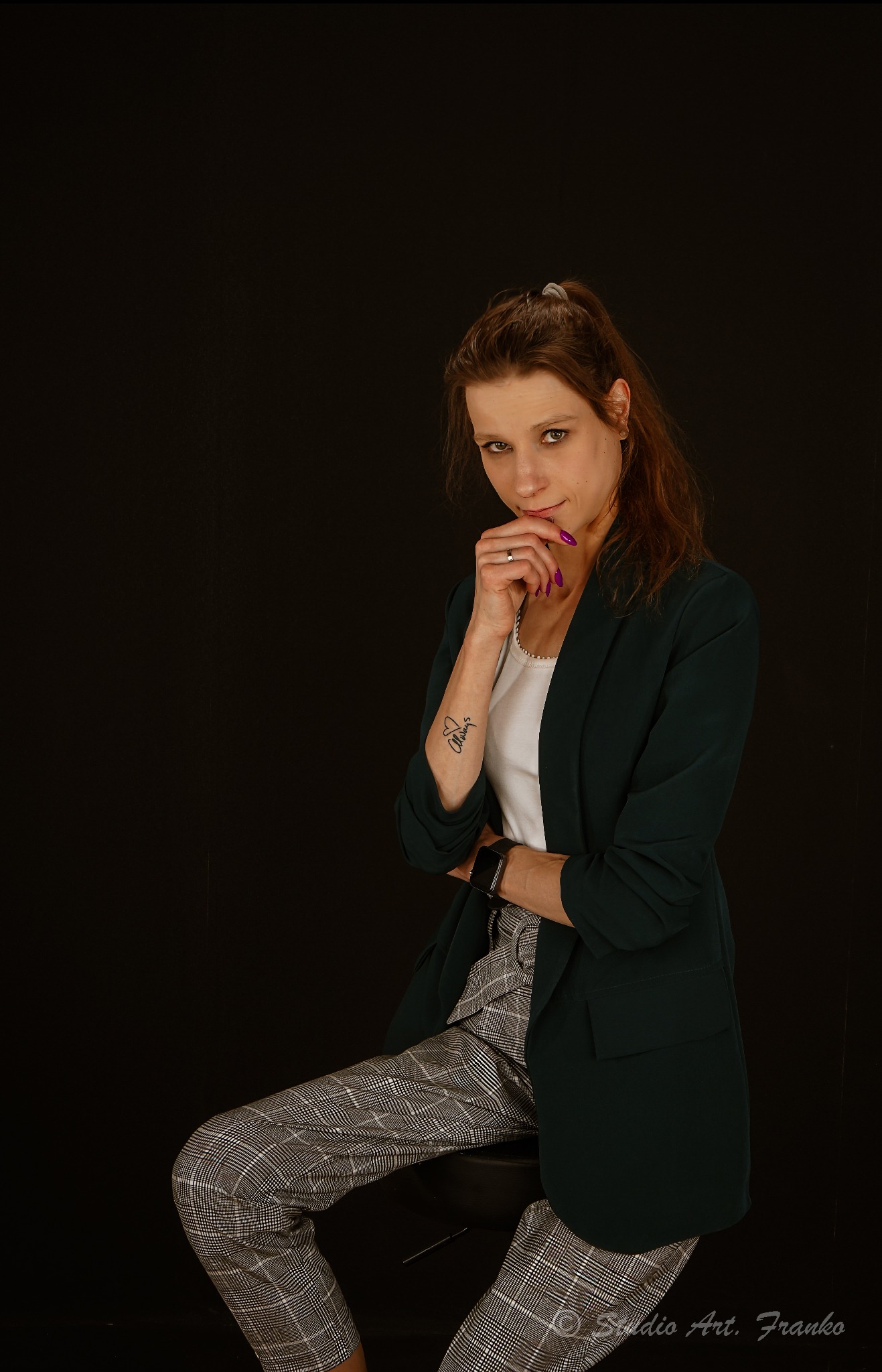 Portret kobiety w zielonej marynarce i kraciastych spodniach na czarnym tle, z tatuażem na ramieniu i pierścionkiem na palcu, siedzącej na krześle.