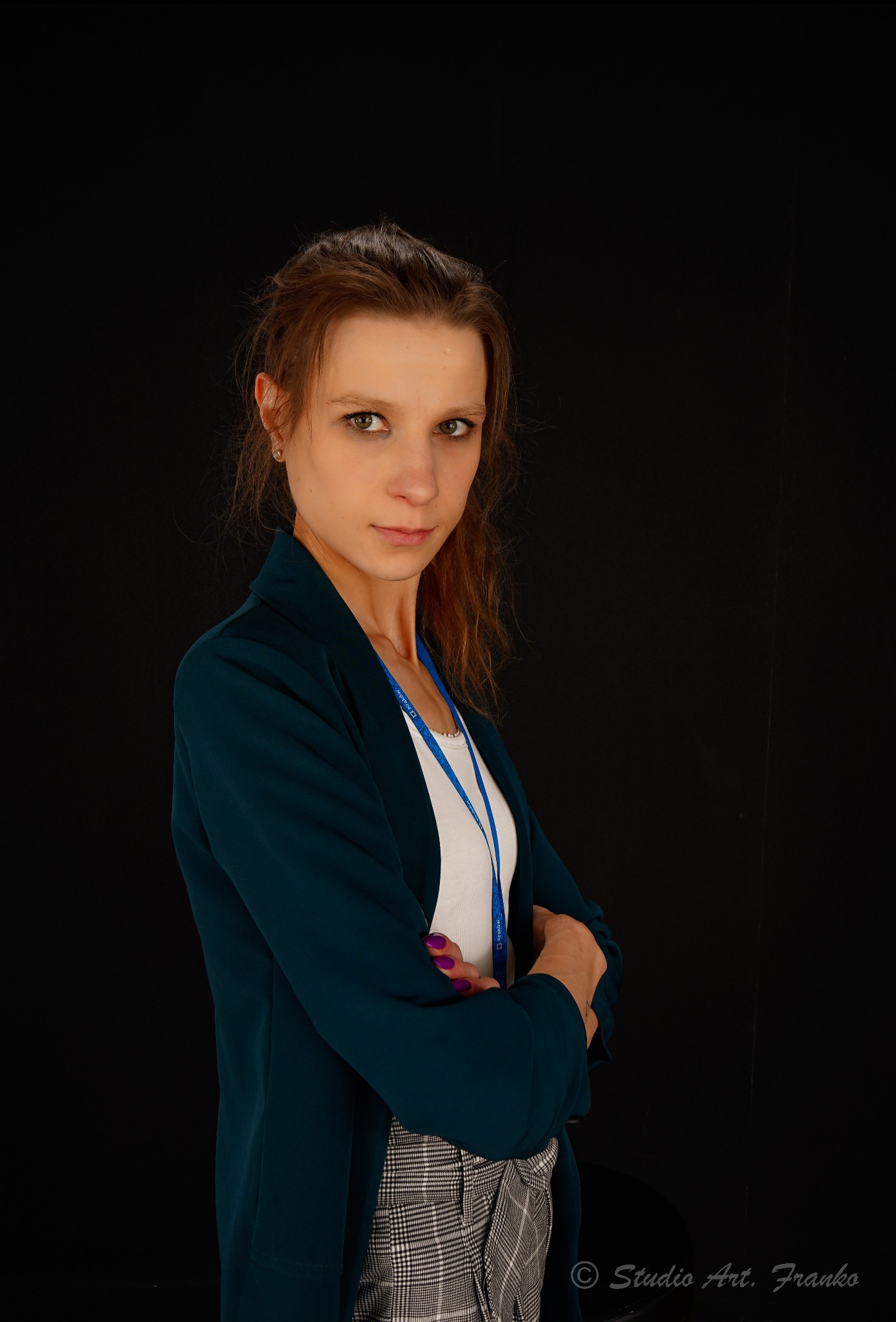 Portret kobiety w ciemnozielonej marynarce i spodniach w kratę na czarnym tle, z rękami skrzyżowanymi, patrzącej wprost w obiektyw, z widocznym identyfikatorem na szyi i fioletowym lakierem...