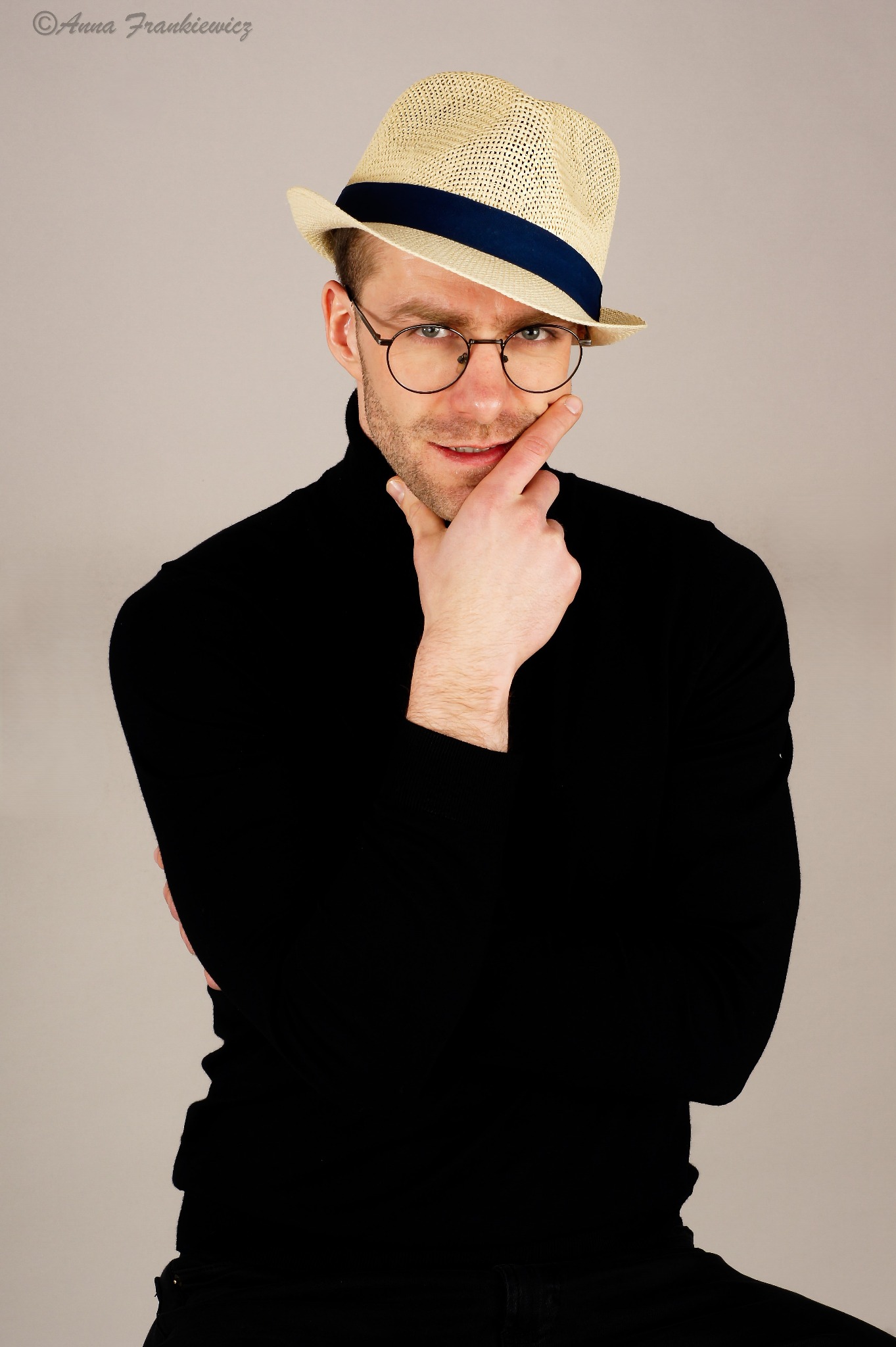 Mężczyzna w czarnym golfie i słomkowym kapeluszu z niebieską wstążką, z okrągłymi okularami, dotyka palcem brody, patrząc wprost.