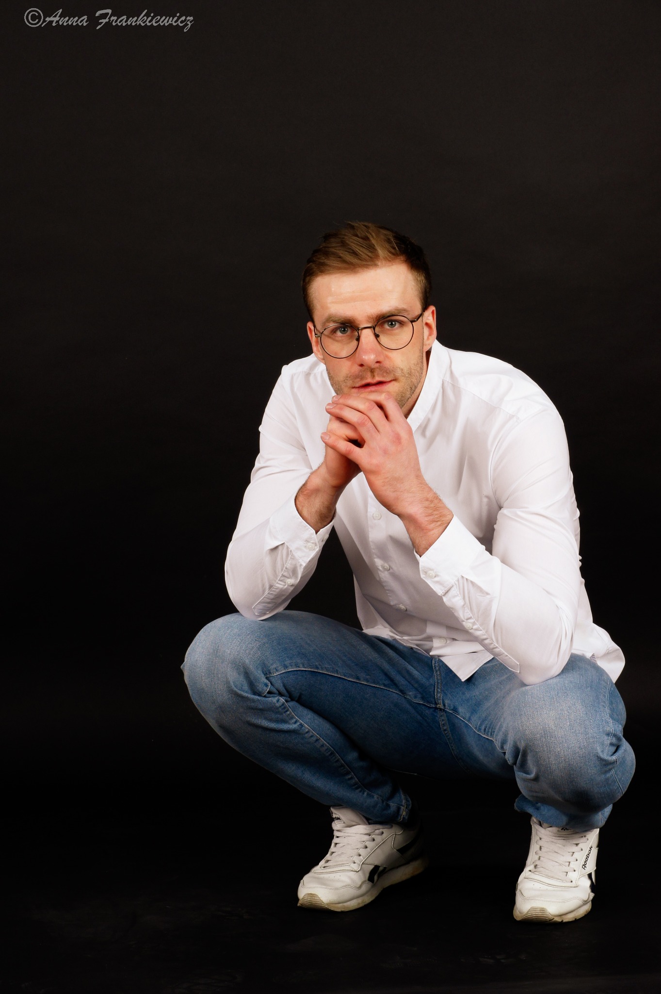 Mężczyzna w okularach, białej koszuli i dżinsach kuca na czarnym tle, dłonie splecione pod brodą, spogląda w obiektyw.