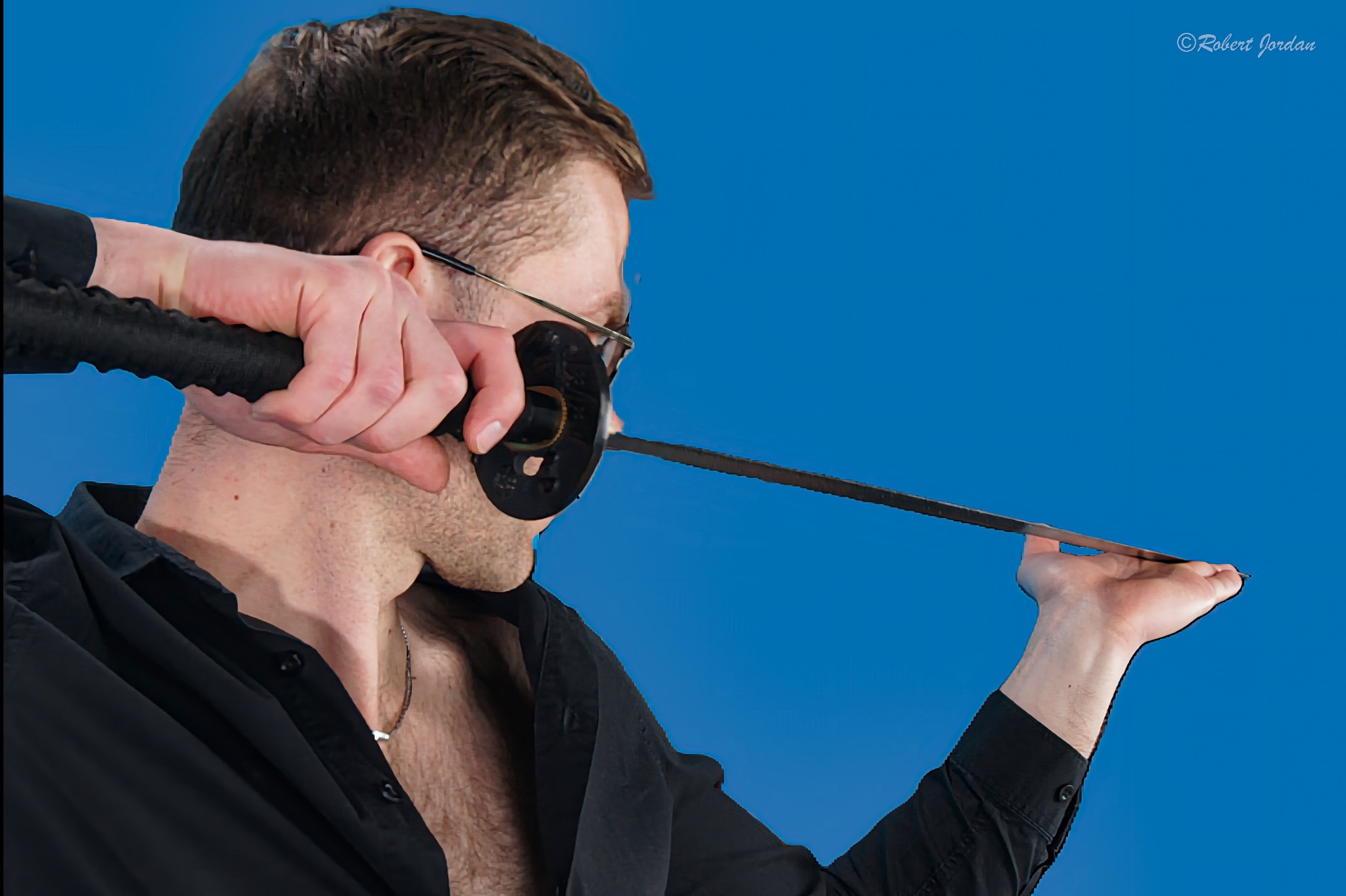 Mężczyzna w czarnej koszuli i okularach demonstruje cięcie mieczem katana na tle błękitnego nieba.