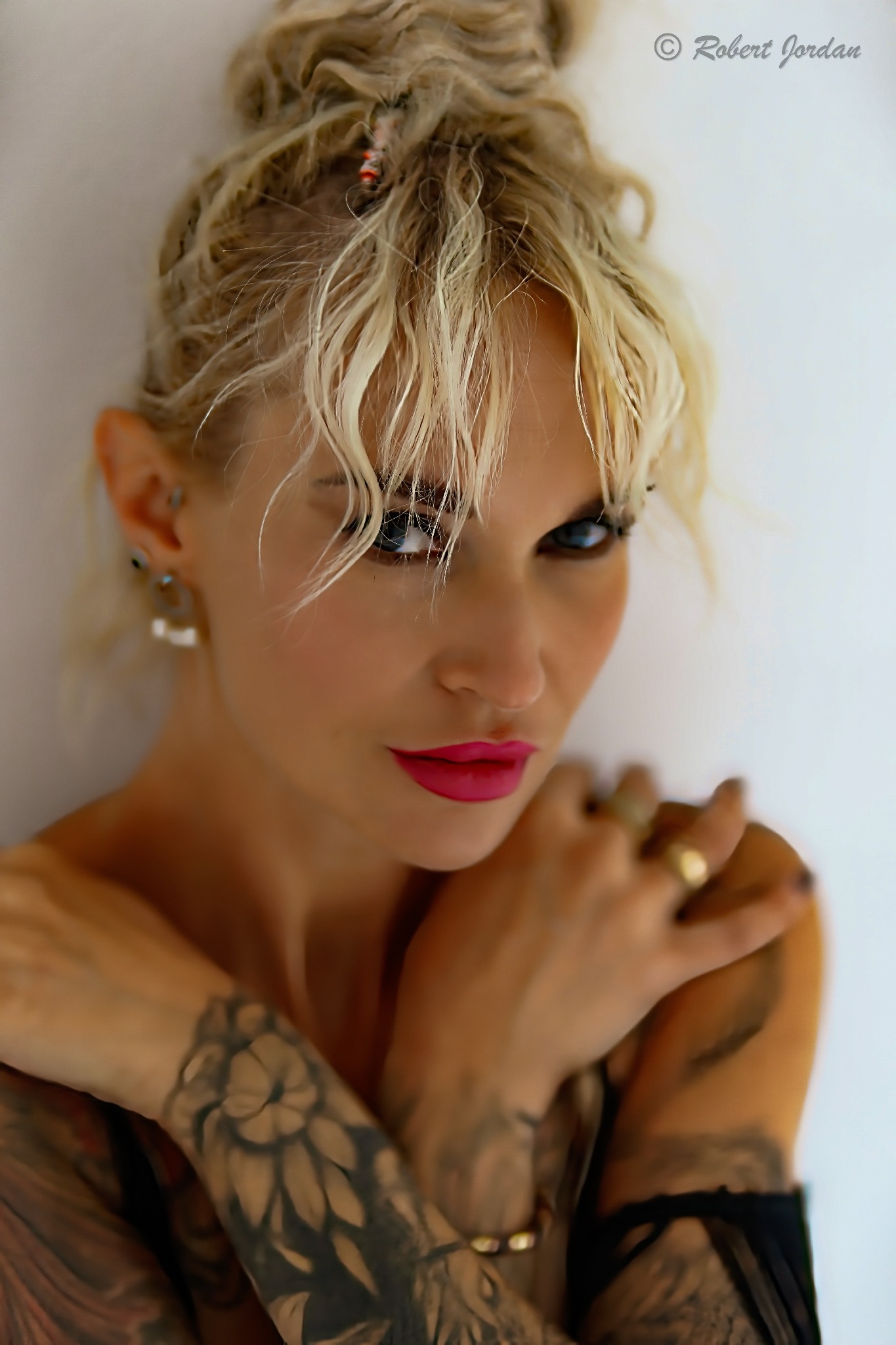 Portret kobiety z blond włosami upiętymi w kok, tatuażami na ramionach i wyrazistym makijażem ust, pozującej na jasnym tle.