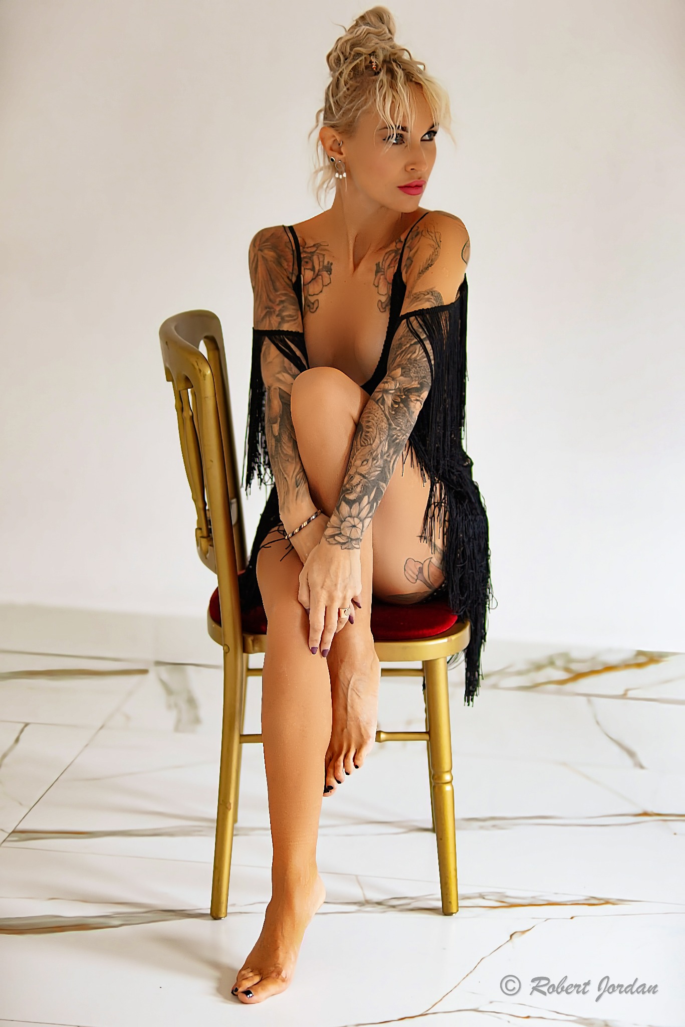 Kobieta z tatuażami siedzi na złotym krześle, ubrana w czarny strój z frędzlami, spięte blond włosy, marmurowa podłoga.