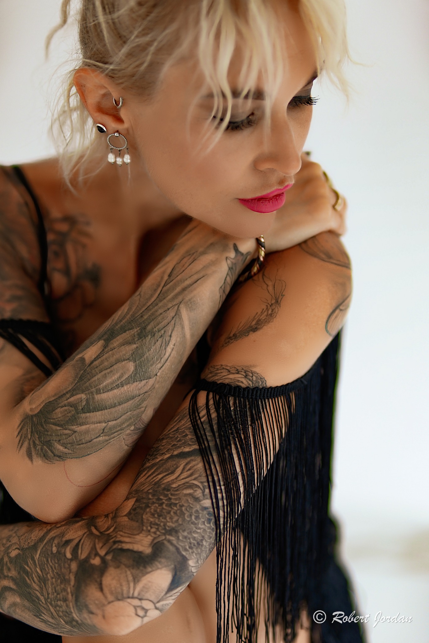 Portret kobiety z platynowymi włosami, ozdobiona tatuażami na ramionach i dekolcie, nosząca kolczyki z perłami i czarną bluzkę z frędzlami.