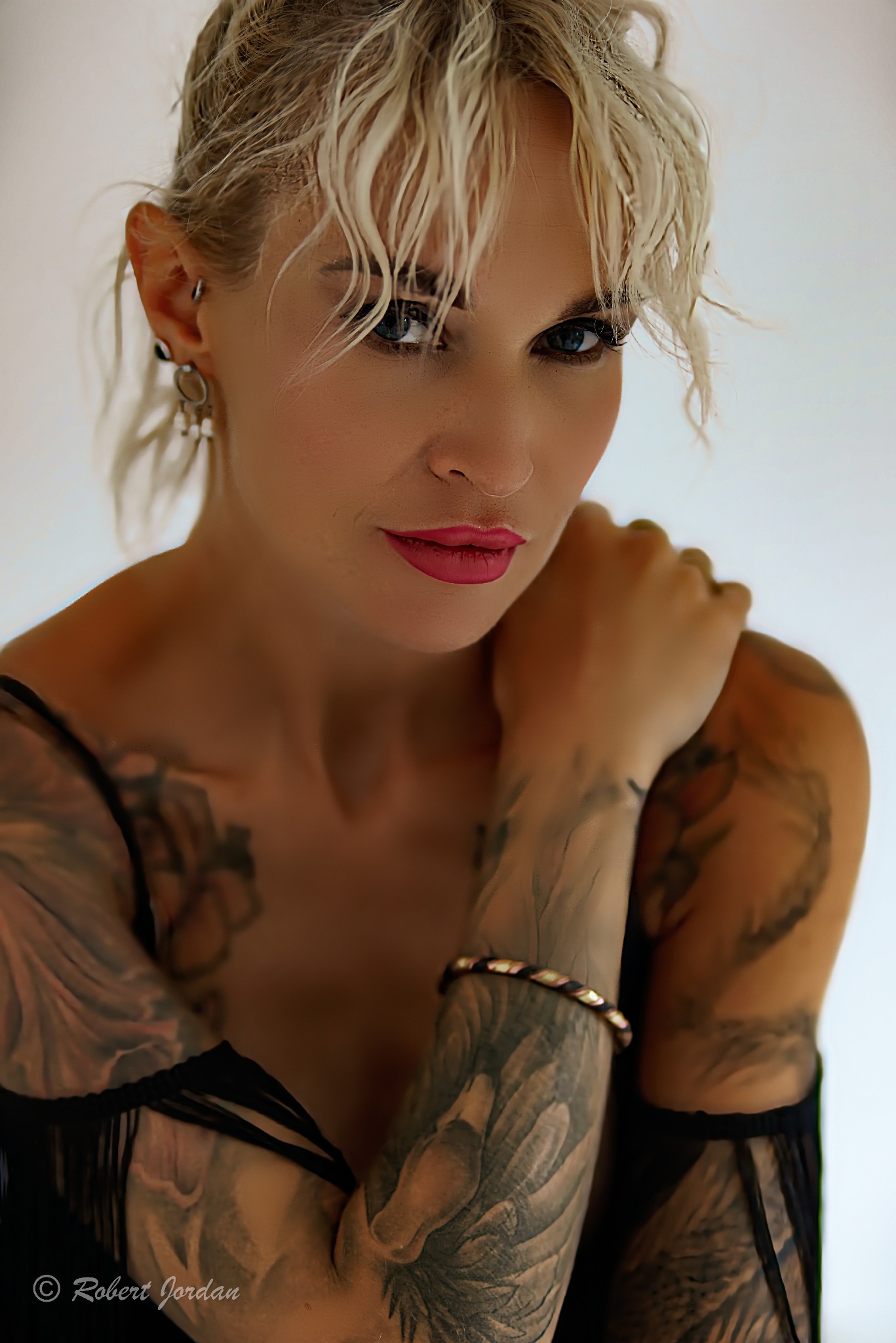 Portret kobiety z tatuażami na ramionach, platynowe blond włosy w nieładzie, intensywne spojrzenie, delikatny makijaż z różowymi ustami, bransoletka na nadgarstku.