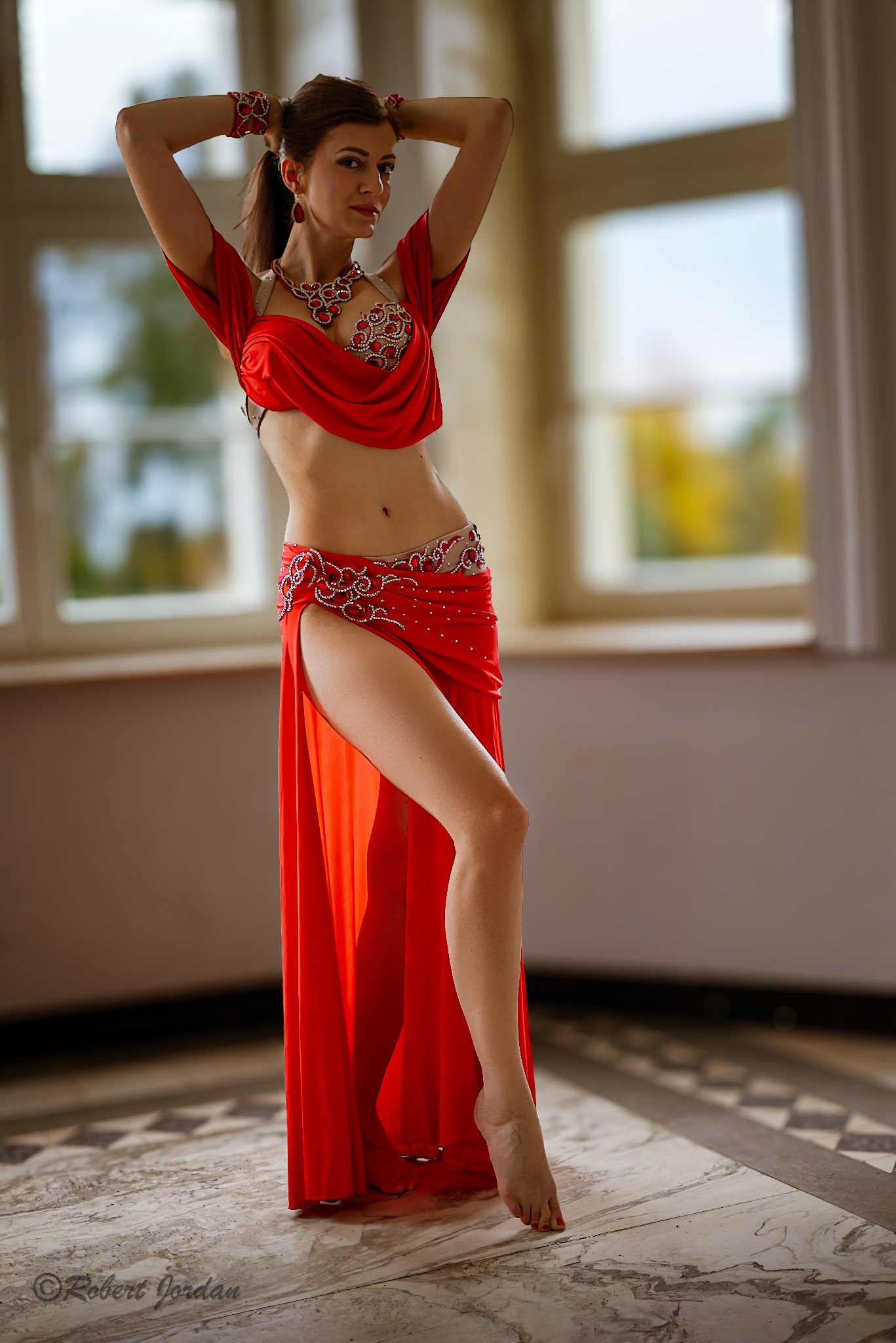 Kobieta w czerwonym, zdobionym stroju do tańca brzucha, pozuje na tle okna w jasnym pomieszczeniu.