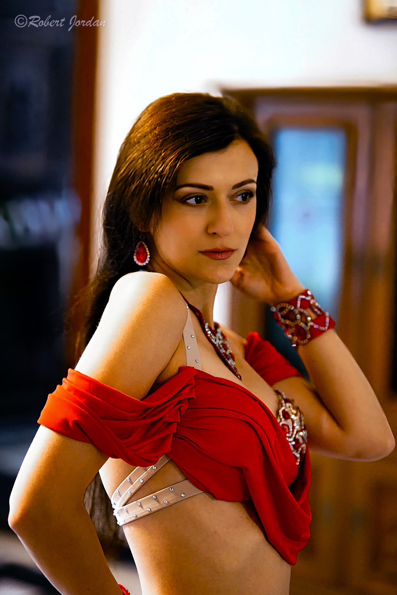 Portret kobiety w czerwonym stroju do tańca brzucha, z bogato zdobionym mankietem i kolczykami, pozującej na rozmytym tle.