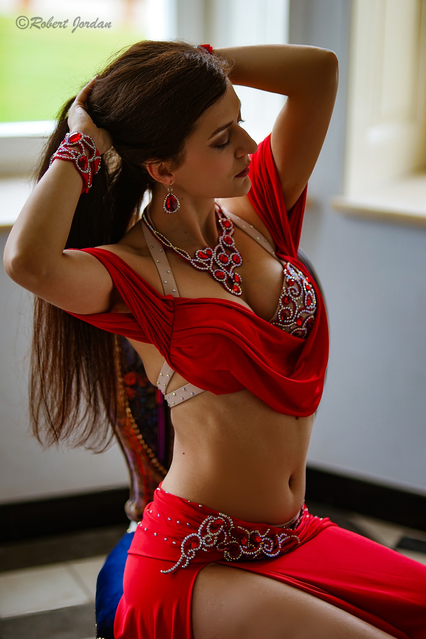 Kobieta w czerwonym stroju do tańca brzucha, z bogato zdobioną biżuterią z czerwonych kamieni, poprawia włosy na tle okna.