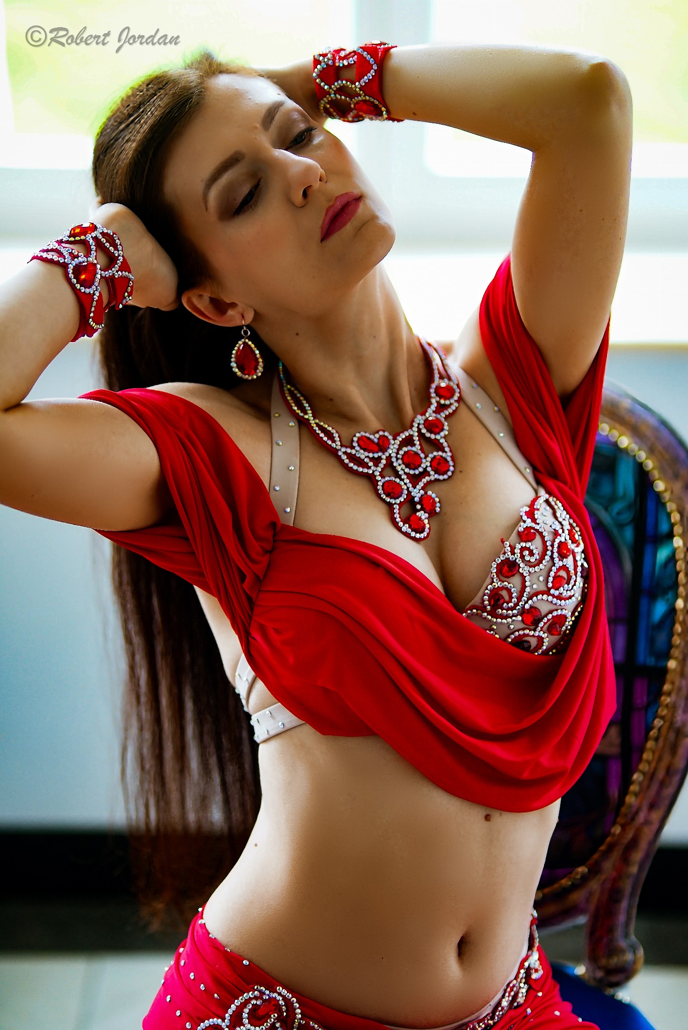 Portret kobiety w czerwonym, bogato zdobionym stroju do tańca brzucha, z rękami uniesionymi nad głową i zamkniętymi oczami, pozująca przy oknie.