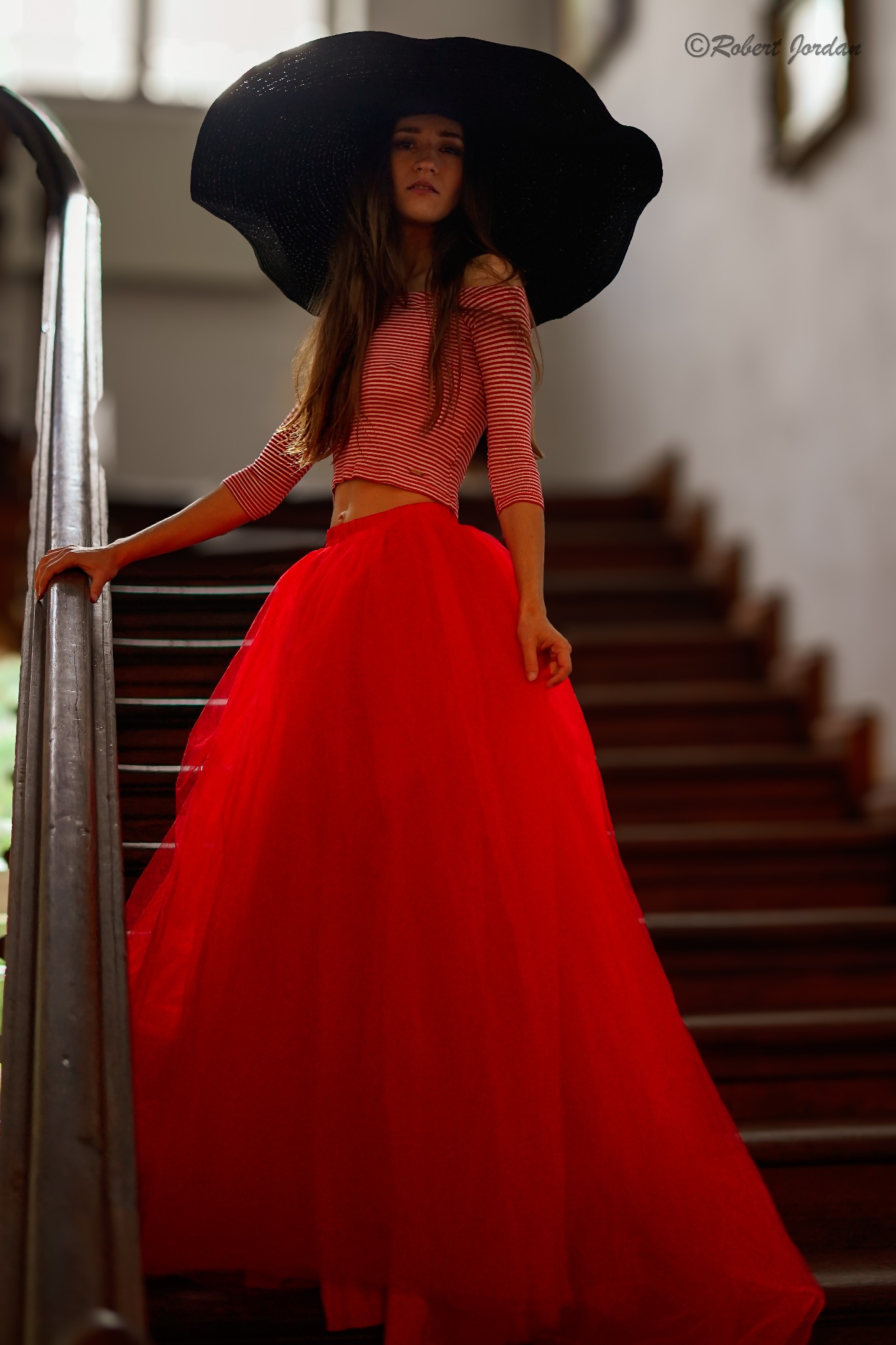 Kobieta na schodach w czerwonej tiulowej spódnicy i kapeluszu z szerokim rondem, pozująca w paski bluzce.