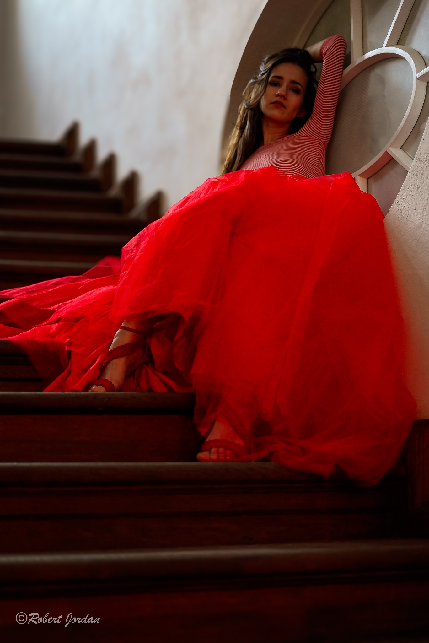 Kobieta w czerwonej, tiulowej spódnicy i bluzce w paski pozuje siedząc na drewnianych schodach, opierając się o okrągłe okno.