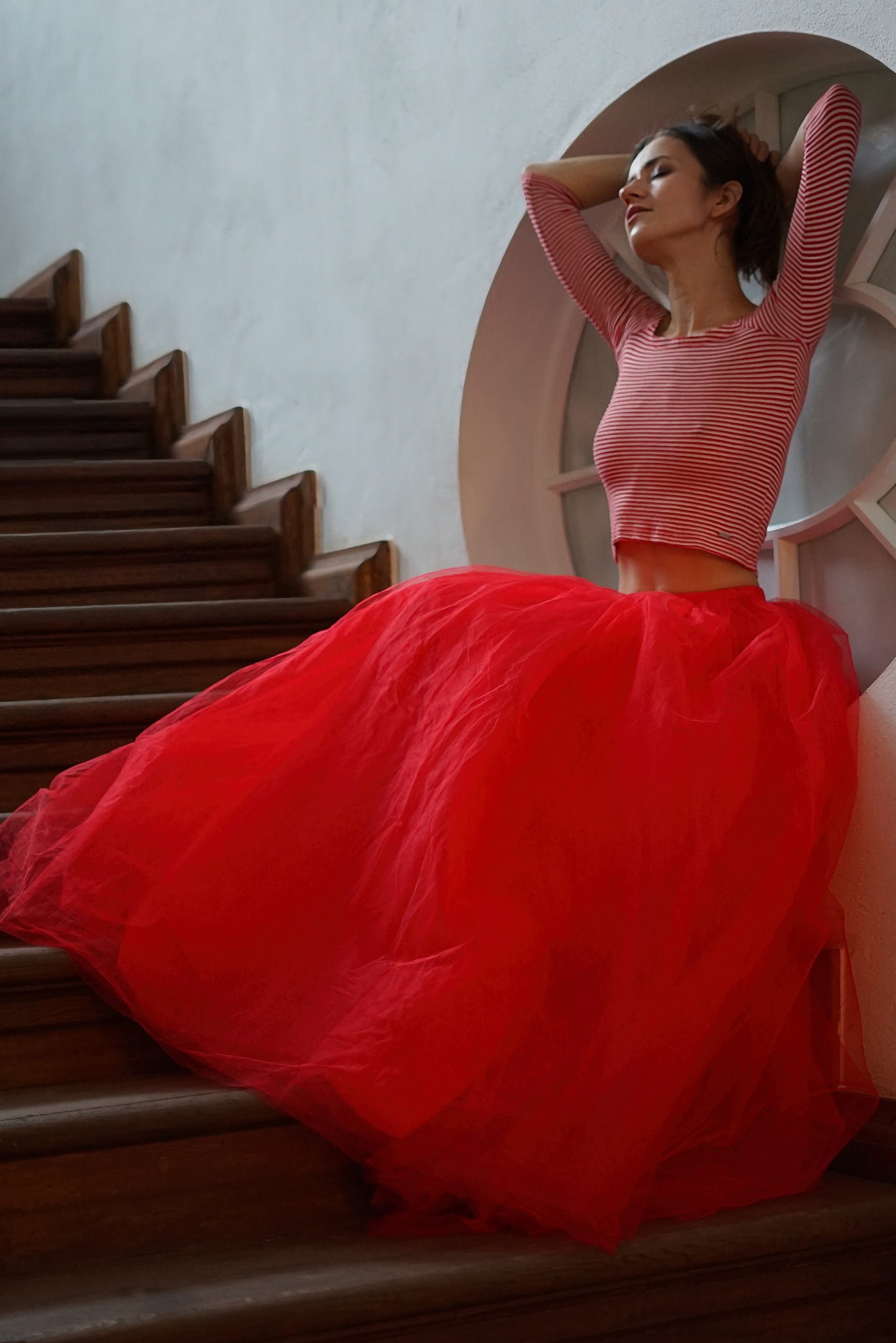 Kobieta w czerwonej tiulowej spódnicy i bluzce w paski pozuje na drewnianych schodach przy okrągłym oknie.