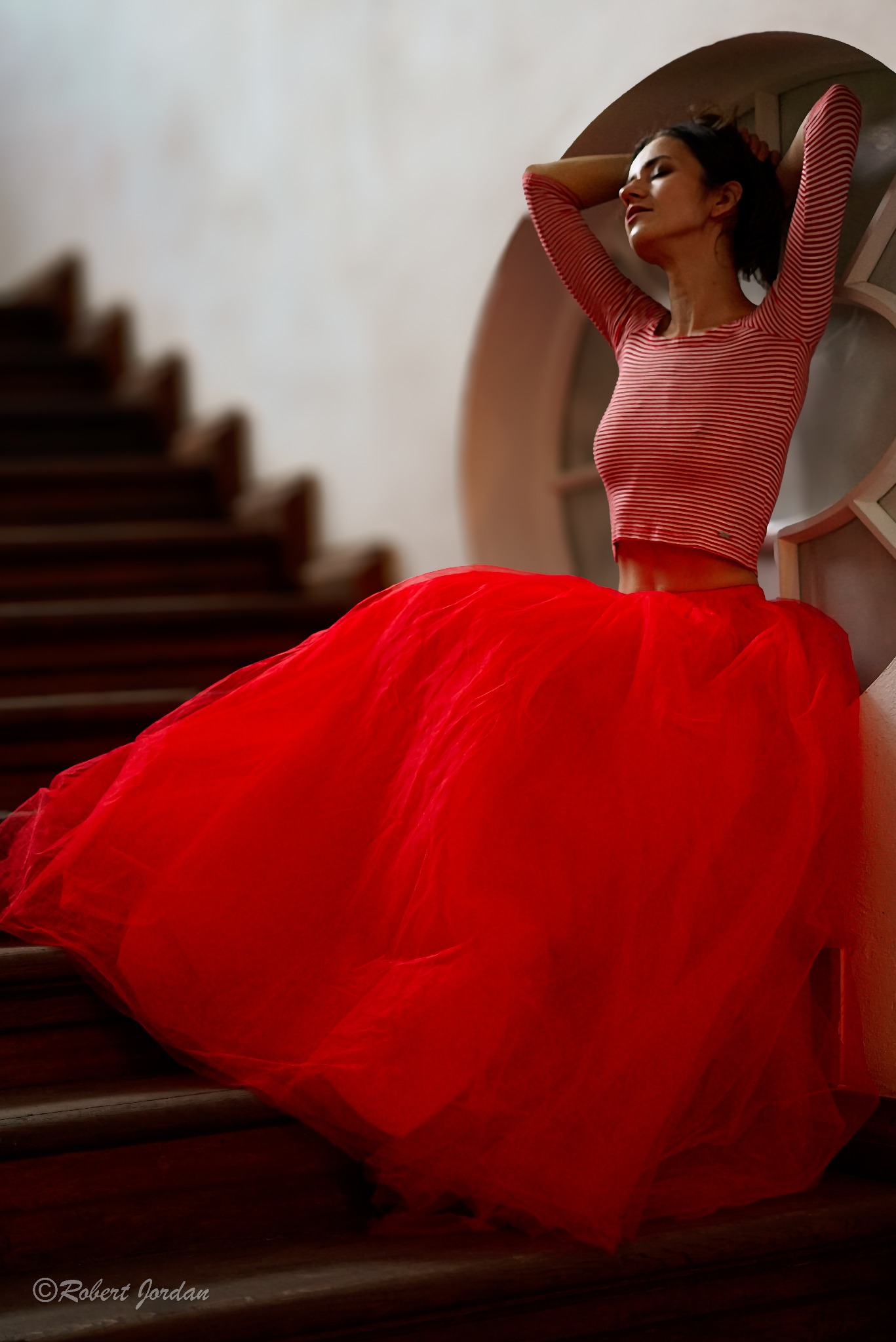 Kobieta w czerwonej tiulowej spódnicy i bluzce w paski pozuje na schodach, ręce uniesione za głową, zrelaksowana poza i zamknięte oczy, okrągłe okno w tle.