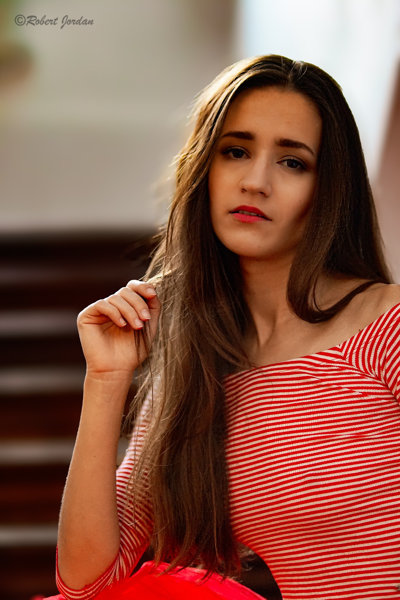 Portret młodej kobiety w bluzce w czerwono-białe paski, z ręką uniesioną do twarzy, w naturalnym oświetleniu.