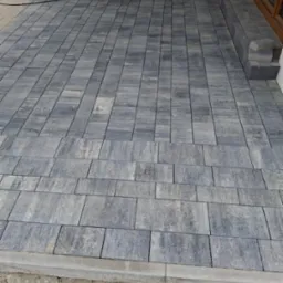 Nowo ułożona, szara kostka brukowa na tarasie, widoczna z góry, z fragmentem okna w tle i wystającą wtyczką elektryczną.