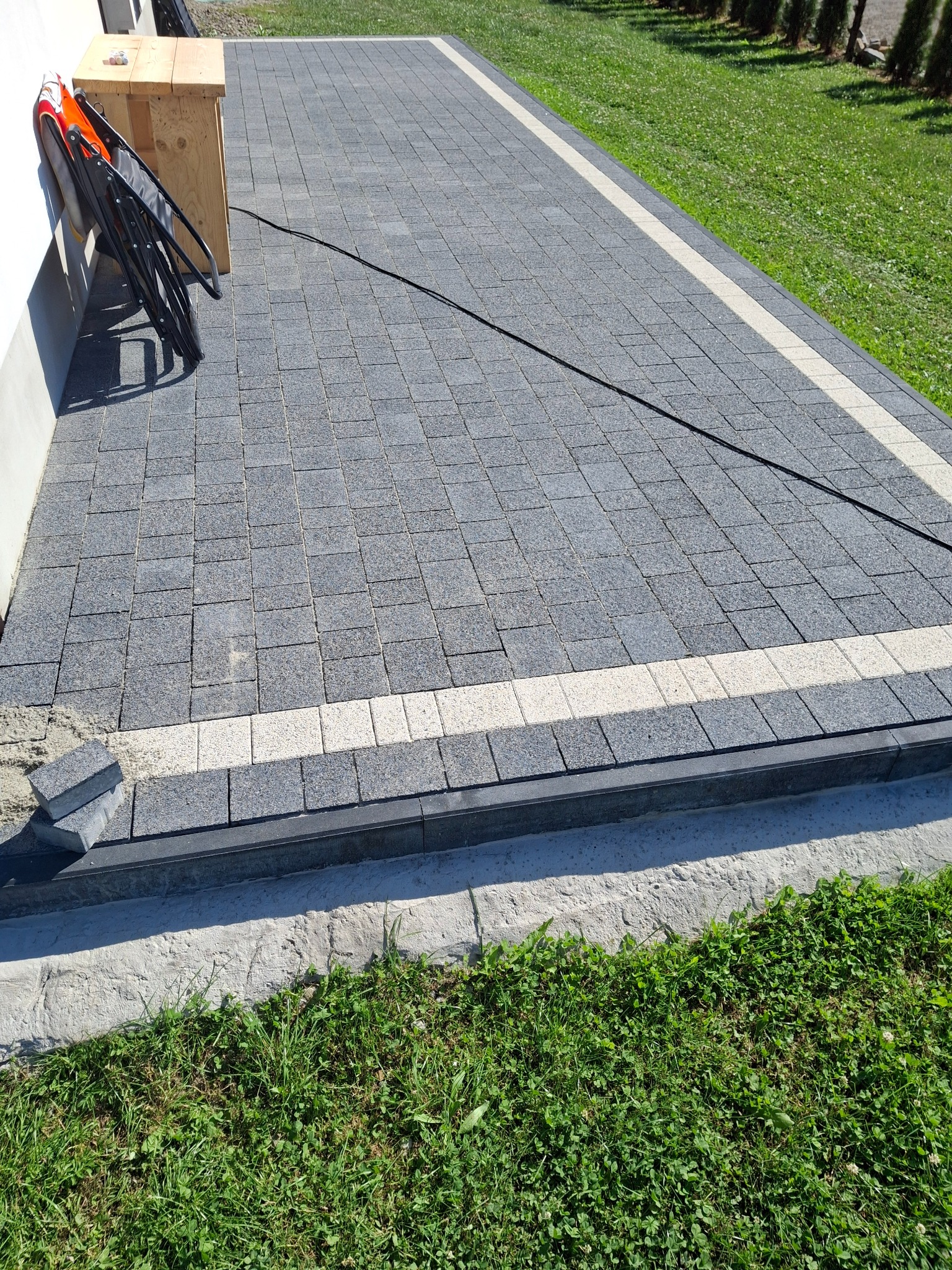 Ułożona szara kostka brukowa z jasnym pasem, widoczne niedokończone krawędzie i zapas kostki, na tle trawnika i budynku.