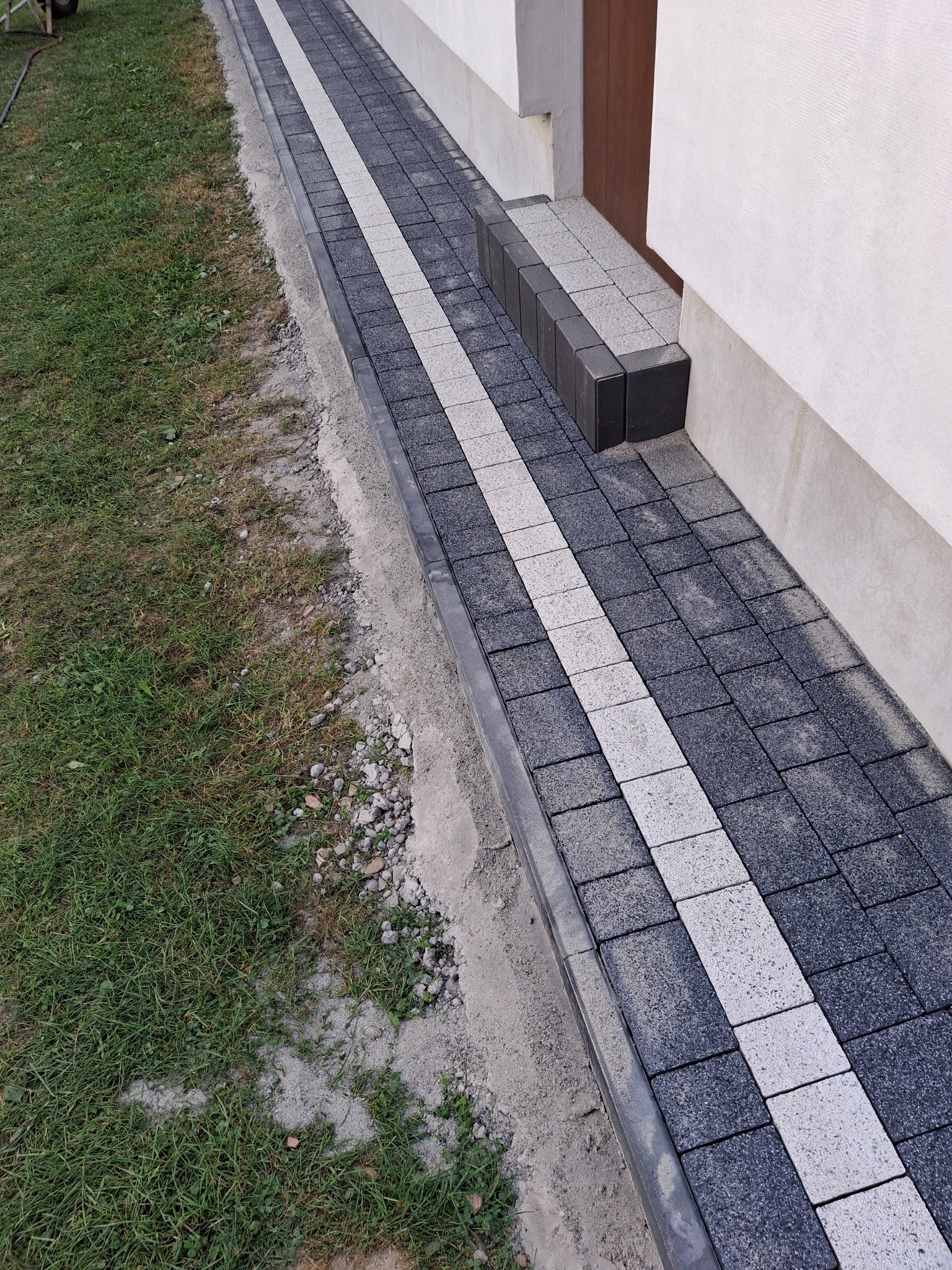 Świeżo ułożony chodnik z ciemnoszarej kostki brukowej z jasnym pasem pośrodku, prowadzący do drzwi wejściowych budynku z trawnikiem po lewej stronie.