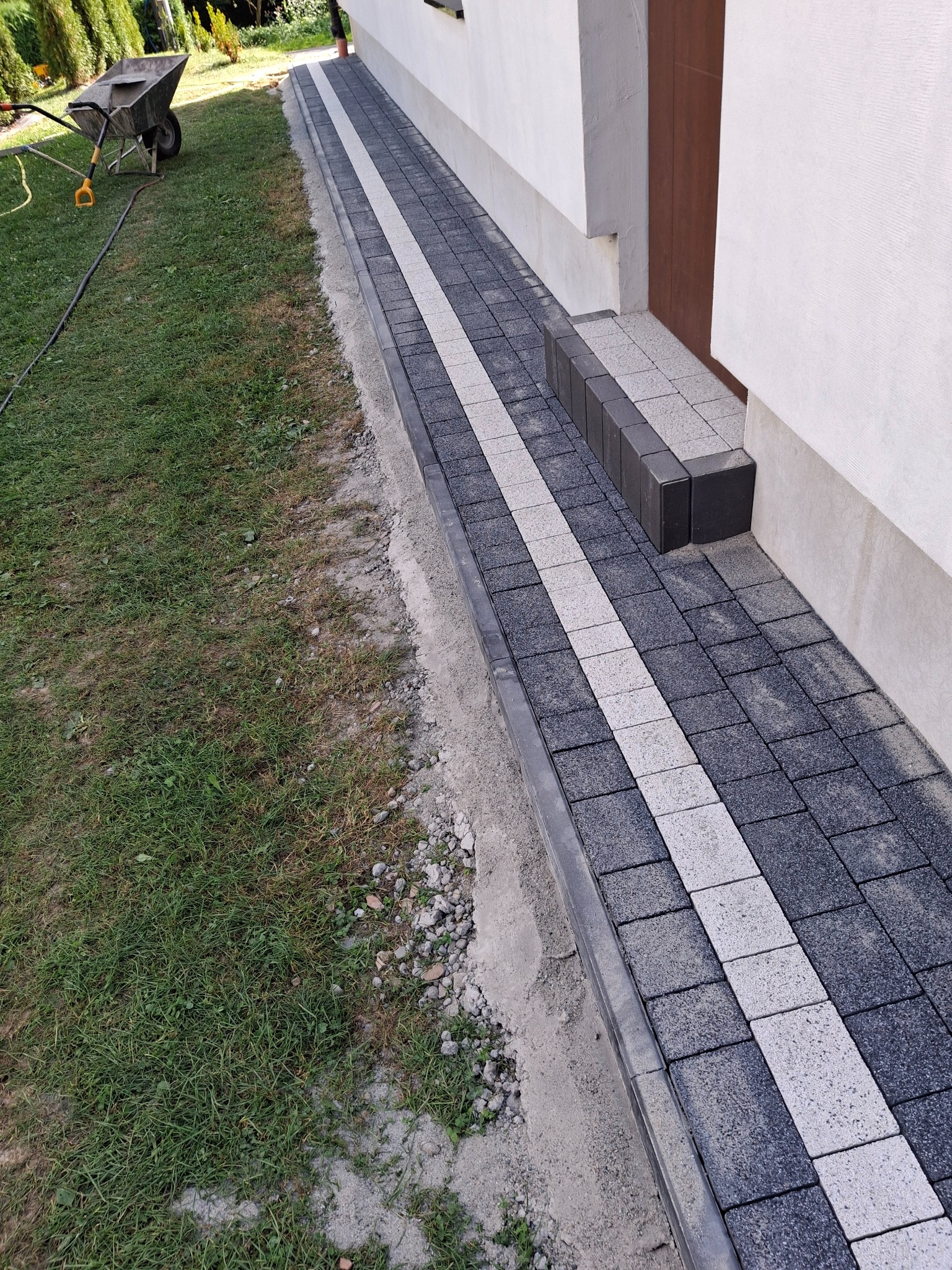Ułożona kostka brukowa w kolorach szarości i bieli tworząca ścieżkę przy budynku z widocznymi schodami z kostki i taczką na trawniku.