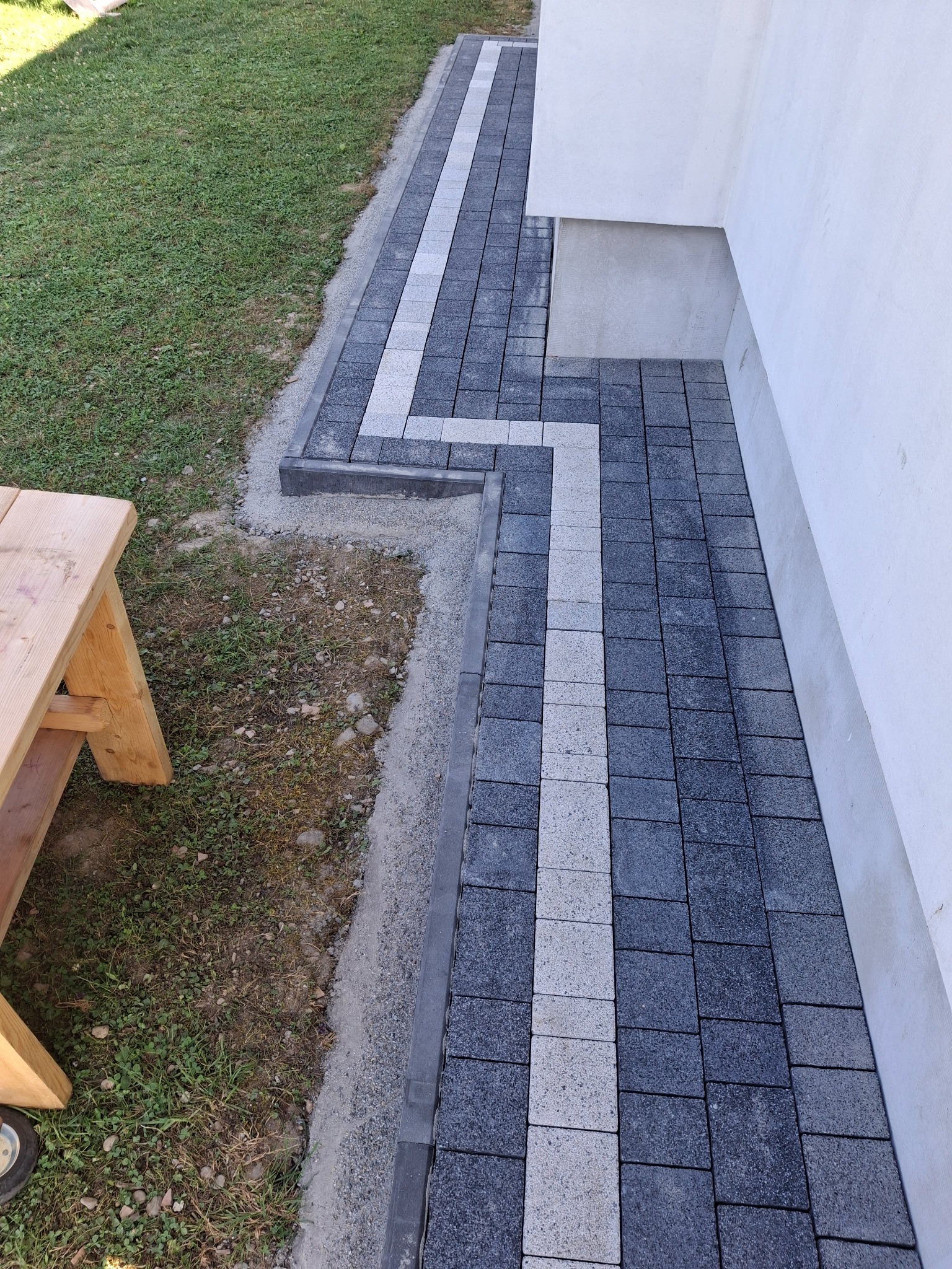 Ścieżka z ciemnoszarej kostki brukowej z jasnoszarym pasem pośrodku, prowadząca wzdłuż białej ściany domu, z widocznym fragmentem trawnika i drewnianego stołka.