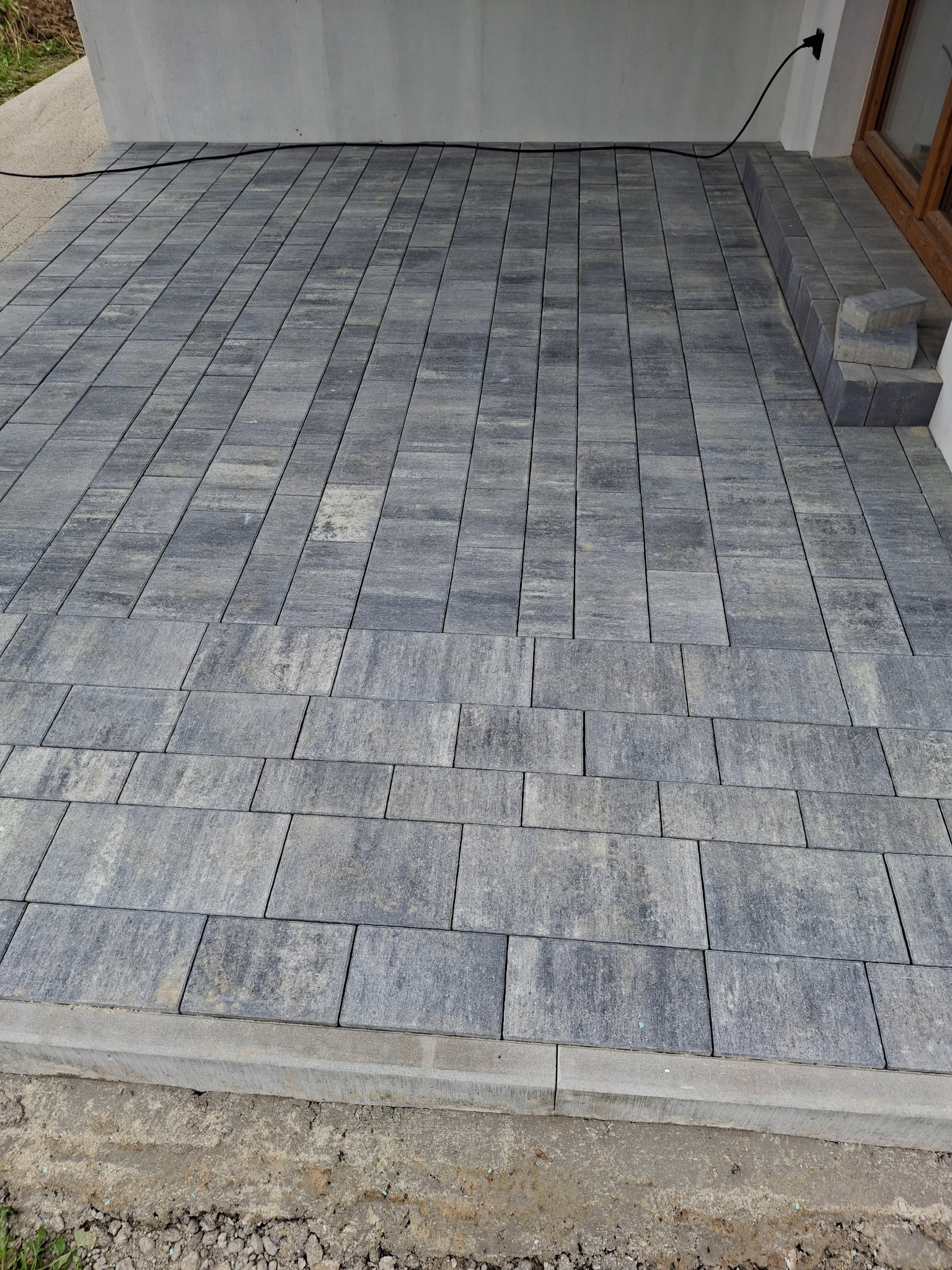 Nowo ułożona, szara kostka brukowa na tarasie, widoczna z góry, z fragmentem okna w tle i wystającą wtyczką elektryczną.