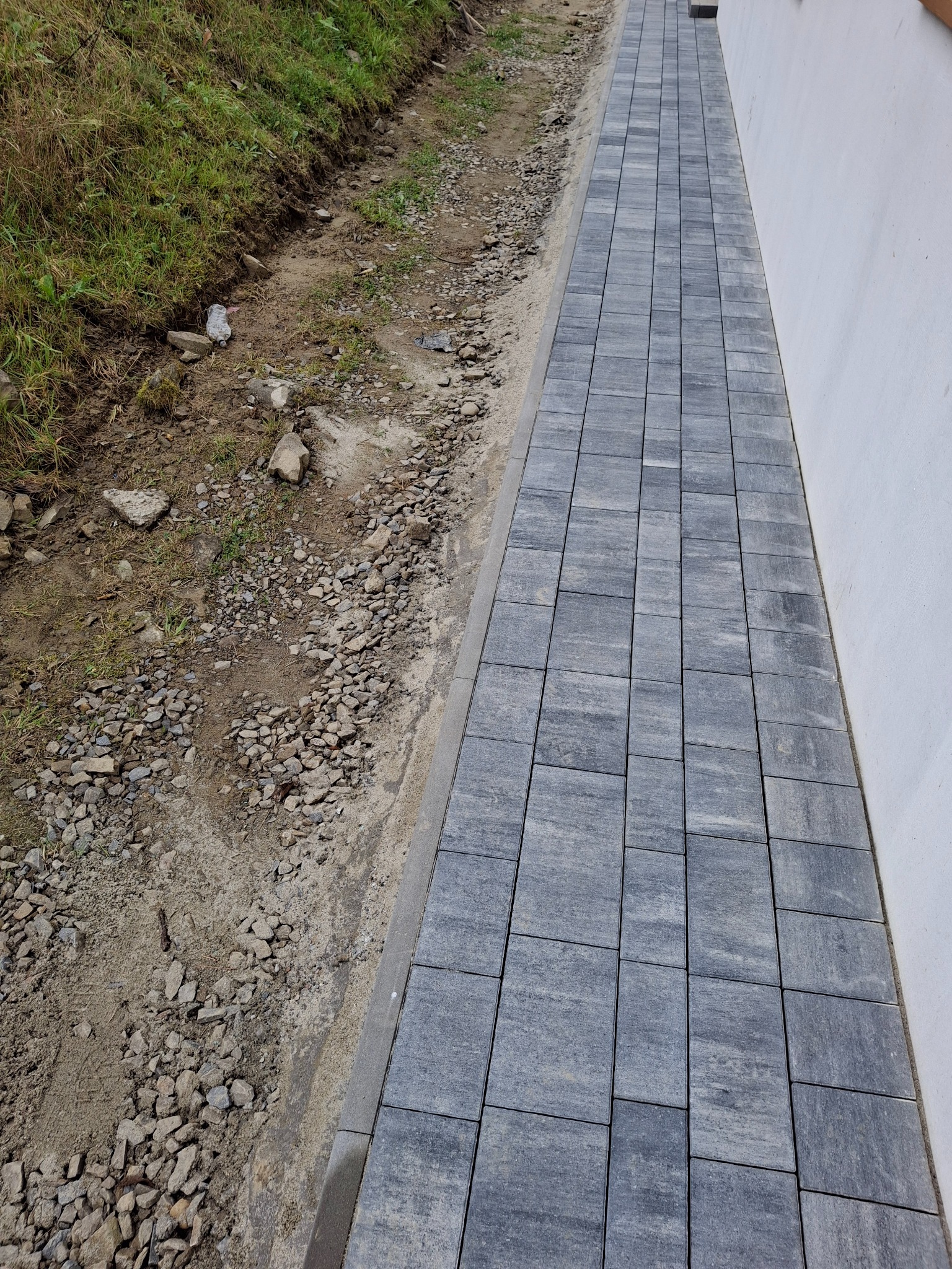 Świeżo ułożona ścieżka z szarych kostek brukowych przylegająca do białej ściany, z widocznym fragmentem ziemi i trawy po lewej stronie.