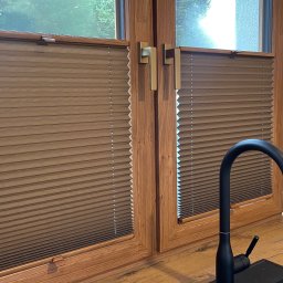 FHU ROL-WIŚ Rolety Wiśniowski - Brązowe, plisowane osłony okienne zamontowane na drewnianych oknach w kuchni, widoczny czarny kran i fragment ściany w trakcie malowania.