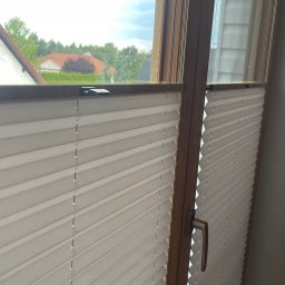 FHU ROL-WIŚ Rolety Wiśniowski - Zamknięte, białe, plisowane osłony okienne w drewnianej ramie, widok z wnętrza na domy w tle.