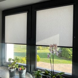 FHU ROL-WIŚ Rolety Wiśniowski - Dwa okna z czarnymi ramami, przesłonięte białymi roletami materiałowymi. Na parapetach stoją doniczki z roślinami, w tym storczyk w fioletowej doniczce. Za oknami widok na zielony ogród.