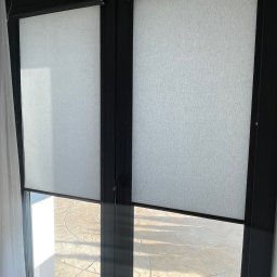 FHU ROL-WIŚ Rolety Wiśniowski - Dwa okna z czarnymi ramami wyposażone w jasne, teksturowane osłony przeciwsłoneczne, częściowo opuszczone, z widokiem na taras z kamienną nawierzchnią.