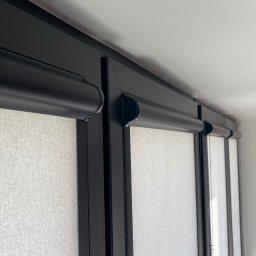 FHU ROL-WIŚ Rolety Wiśniowski - Fragment okna z zamontowanymi jasnymi roletami w czarnych kasetach, widok z dołu, z boku zwisająca biała firanka.