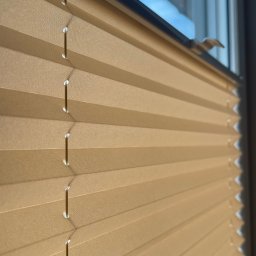 FHU ROL-WIŚ Rolety Wiśniowski - Beżowe, plisowane osłony okienne w pozycji zamkniętej, widoczne sznurki i fragment okna z odbijającym się niebem.