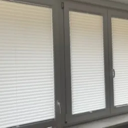 Trzy okna z zamontowanymi białymi plisami harmonijkowymi, widoczne detale mechanizmów sterujących.