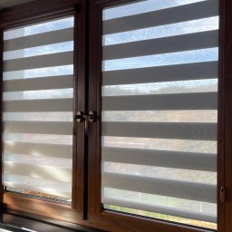 FHU ROL-WIŚ Rolety Wiśniowski - Dwa okna z drewnianą ramą i zamontowanymi roletami w systemie dzień i noc, z widokiem na zewnątrz przez prześwity.