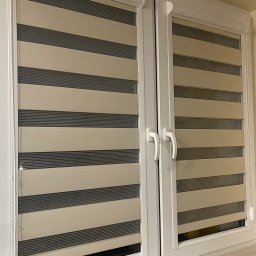 FHU ROL-WIŚ Rolety Wiśniowski - Dwuskrzydłowe okno z zamontowanymi roletami w biało-szare pasy, widoczne białe ramy okienne i parapet z jasnego kamienia.