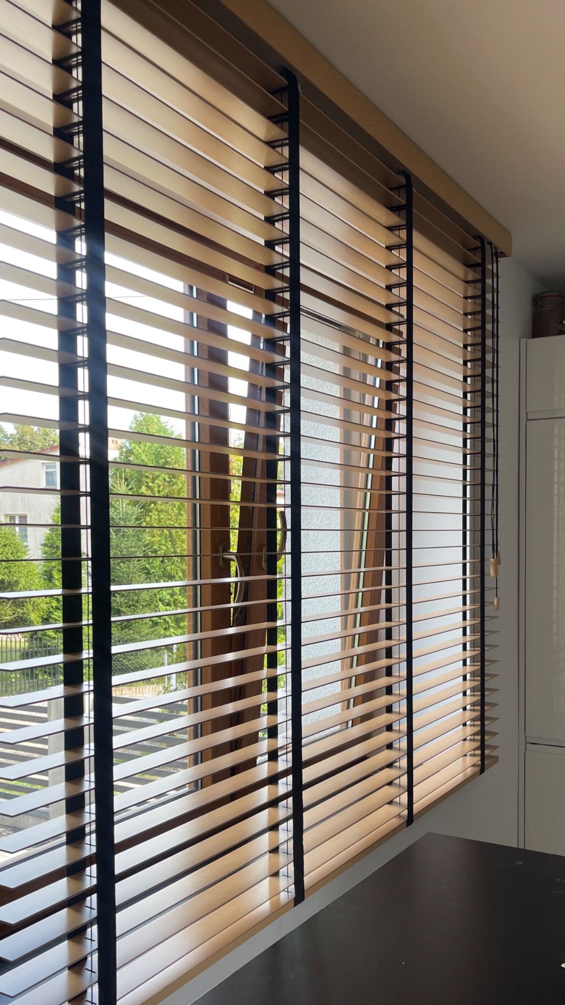 Jasne, drewniane żaluzje poziome z czarnymi taśmami, częściowo odsłonięte, widok na zielone drzewa za oknem.