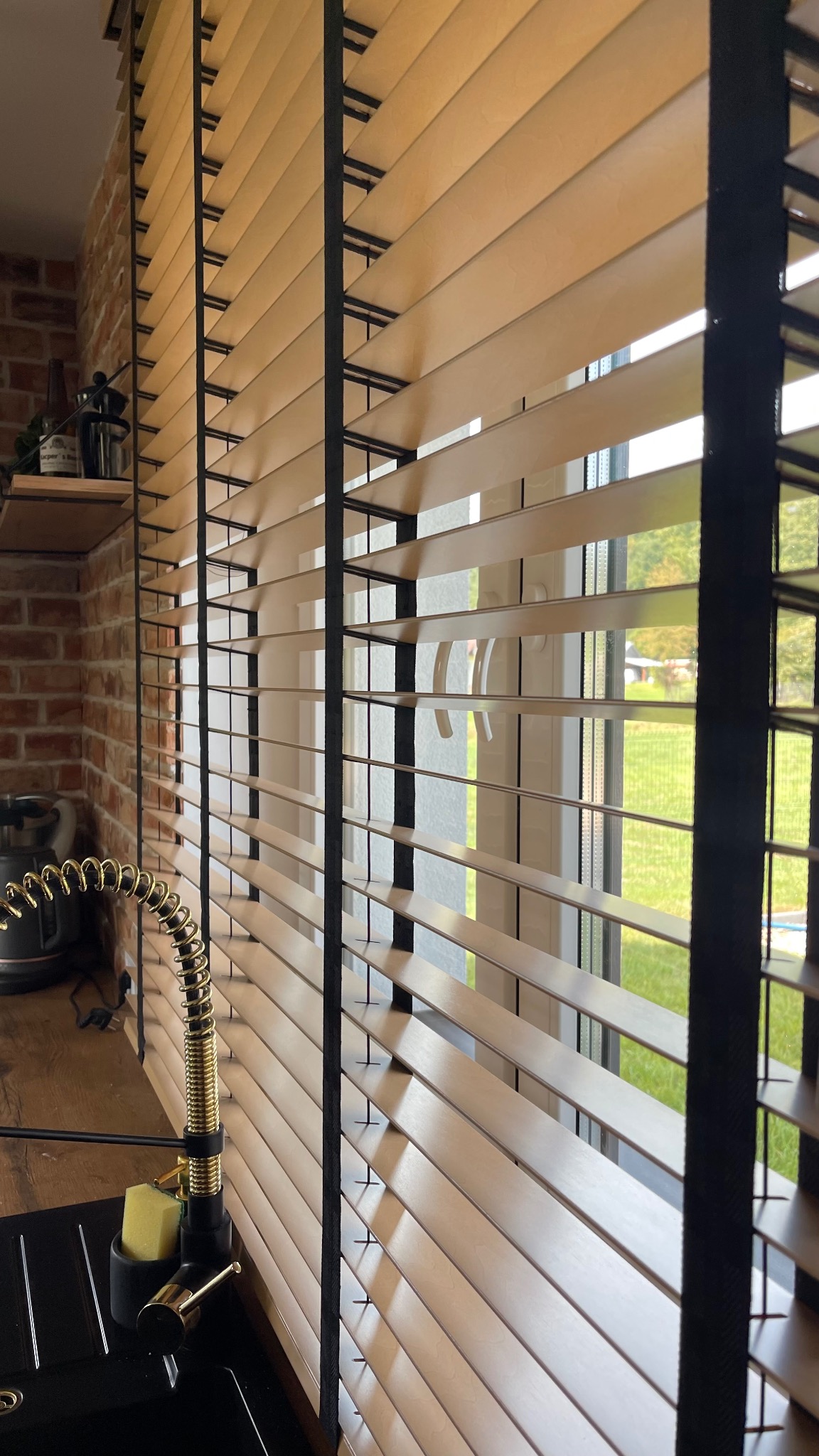 Jasne, drewniane lamele z czarnymi taśmami w oknie kuchennym, widoczny fragment zlewu z baterią i ceglana ściana w tle.