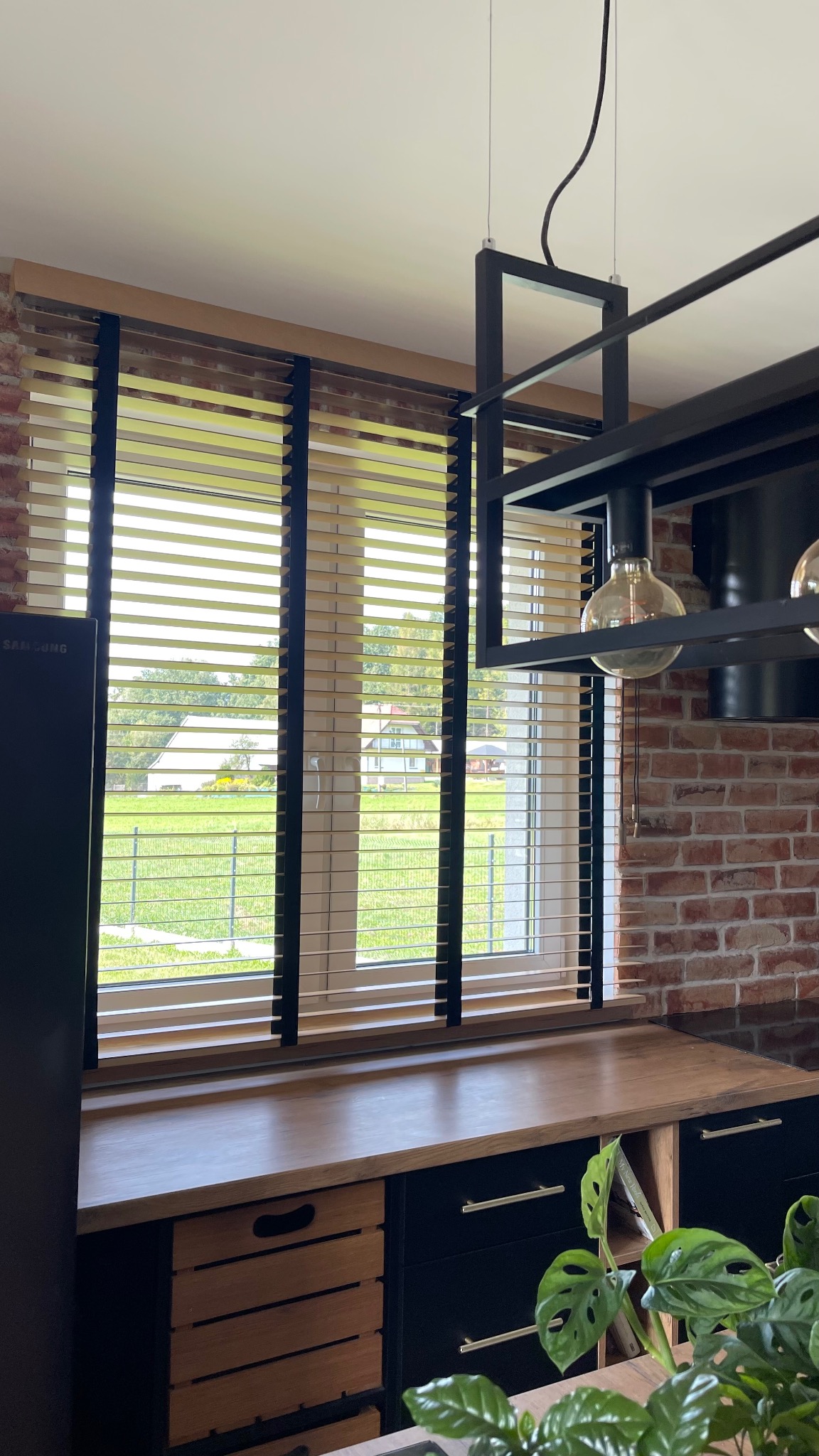Jasne drewniane żaluzje z czarnymi taśmami na oknie z widokiem na zielony ogród, w tle ceglana ściana i nowoczesna lampa wisząca.