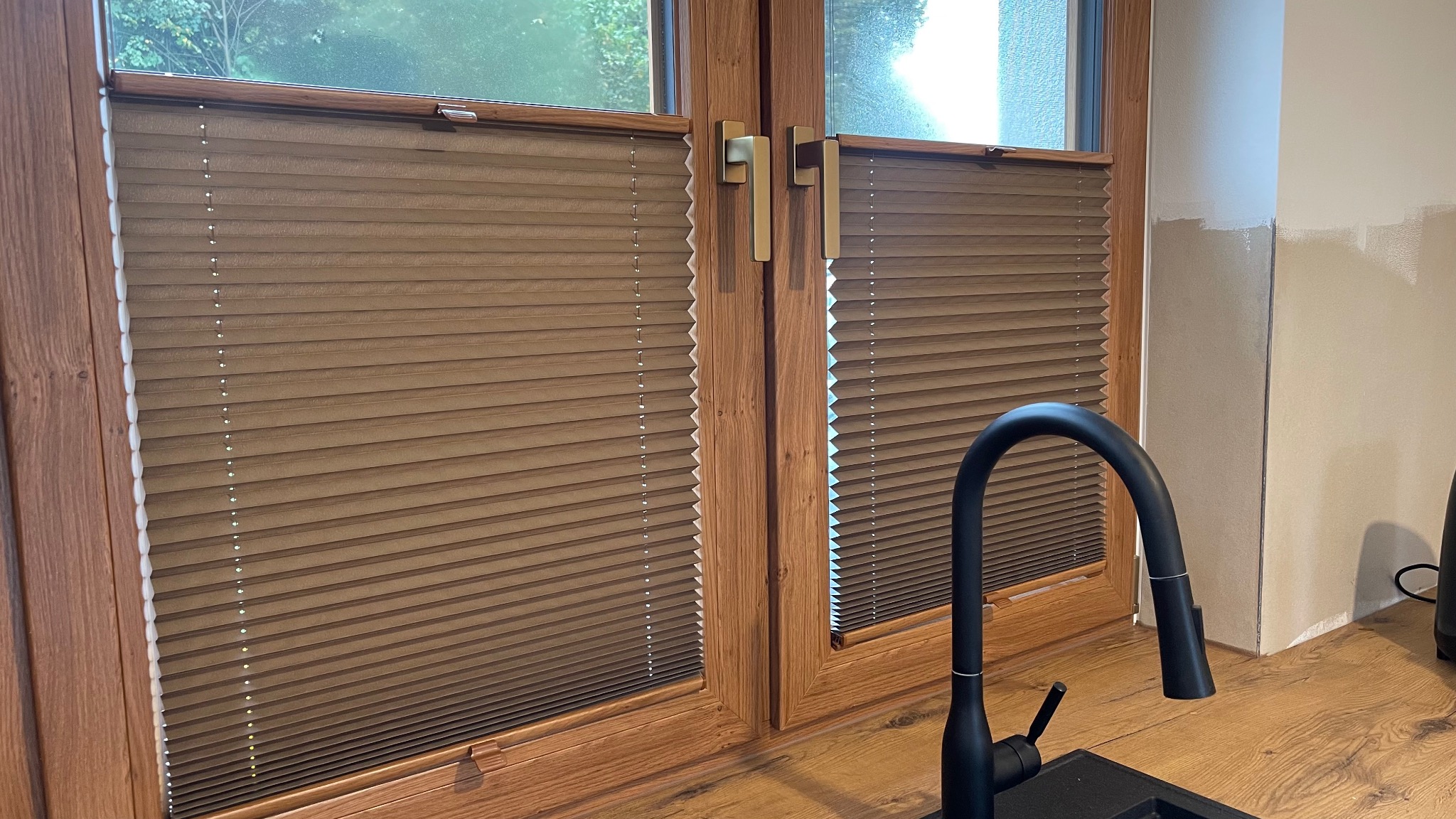 Brązowe, plisowane osłony okienne zamontowane na drewnianych oknach w kuchni, widoczny czarny kran i fragment ściany w trakcie malowania.